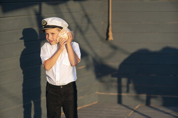 Little ship captain on the deck of a pirate ship. The boy stands on the deck of the ship with a shell in his hands. The boy listens to the sea shell.