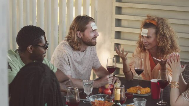 Waist up slowmo of group of young diverse cheerful friends sitting around table in summerhouse playing guessing game and drinking red wine