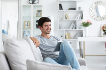 Handsome smiling man is using digital tablet in casual wear on sofa in living room. Remote communication, online shopping
