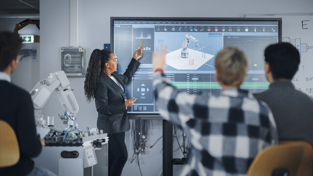 Caucasian Student Girl Raise Her Hand and Asking Teacher Standing Near the Big Screen. Teenagers Studying Robotic Arm at the Lesson In Modern University of Technology. Machine Learning Concept
