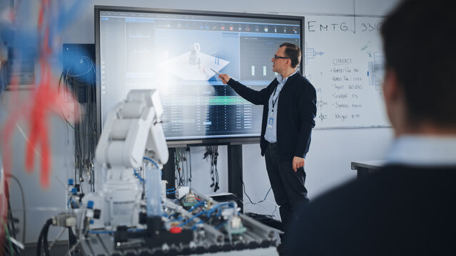 Male Professor Showing at the Big Screen During the Lesson. Diverse Group of Students Working Together on Modification of Functions of Technology Robot Arm for Engineering Project at University
