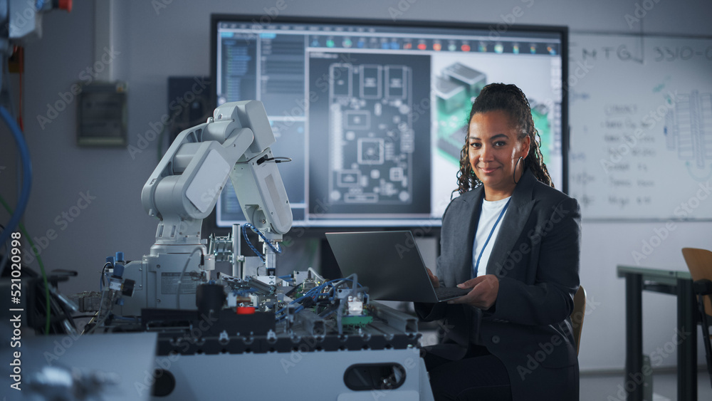 Poster Portrait of Smiling Black Developer Using Laptop while Working with Robotic Arm in Modern University Classroom. Woman Looking at Camera. Success and Programming Concept. Medium Shot