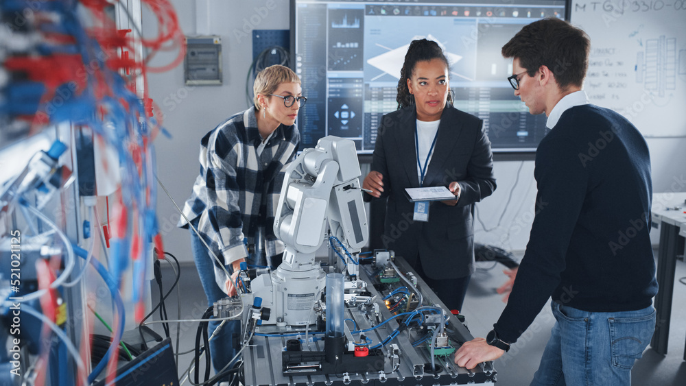 Poster In Robotics Development Laboratory: Black Female Teacher and Two Students Work With Prototype of Robotic Hand. Young Student Telling Something with Smile and Brainstorming with Her Team.