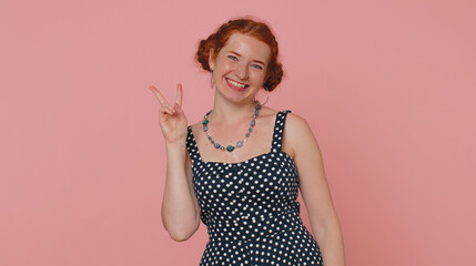 Redhead young woman in polkadot dress showing victory V sign, hoping for success and win, doing peace gesture, smiling with kind optimistic expression. Ginger girl indoors isolated on pink background
