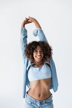 excited african american woman in blue denim shirt standing with raised hands isolated on grey