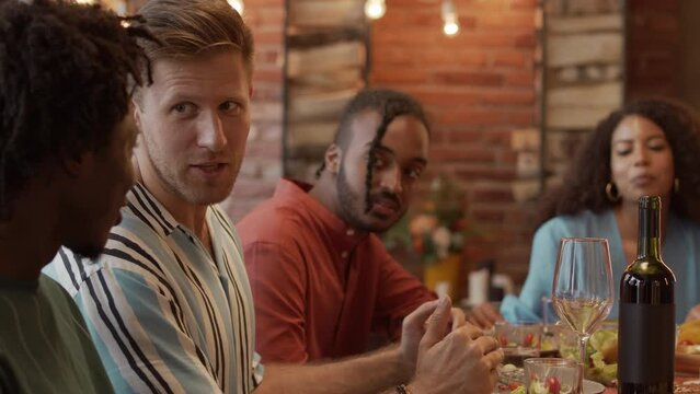 Waist up slowmo of young fancy diverse company of friends having conversation at festively decorated dinner table indoors