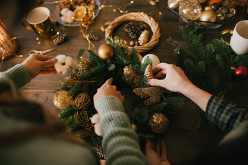 Two millennial women making Christmas wreath using pine branches and festive decorations. Small...