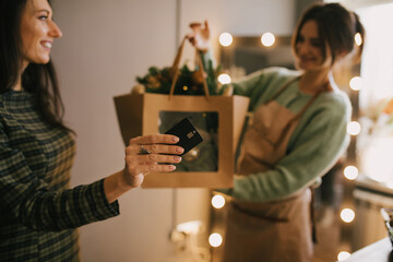 Millennial woman buying Christmas wreath in a cozy flower shop using credit card.