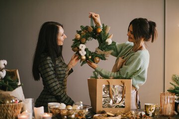 Two millennial women making Christmas wreath using fresh pine branches and festive decorations.