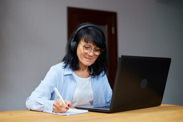 Senior woman wearing wireless headphones watch webinar, trainning, course, lessons on laptop