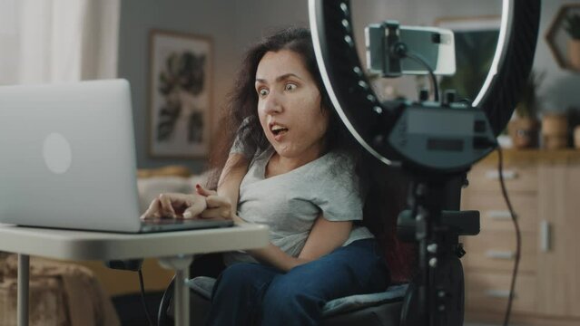 Woman with a spinal muscular atrophy speaking and sitting in a motorized wheelchair at the table with laptop, while shooting a video on smartphone on a tripod with ring LED lamp