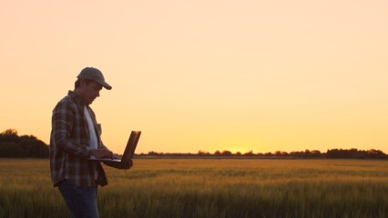 Farmer with a laptop computer in front of a sunset agricultural landscape. Man in a countryside...