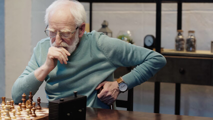 senior man in eyeglasses thinking near chessboard at home