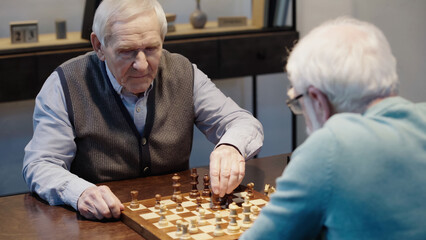 grey haired man playing chess with blurred friend in living room