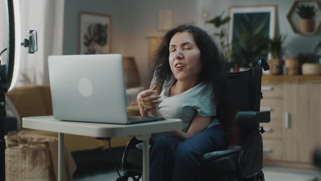 Woman with disability sitting in a wheelchair at home in a cozy room recording a video blog or making a stream in social networks, using a laptop and smartphone on a tripod with a round LED backlight