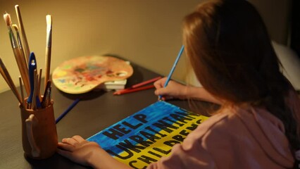 Shooting over shoulder of cute little girl painting Help Ukrainian children placard sitting at table in bomb shelter. Blurred child drawing banner with national flag colors on air-raid alert warning