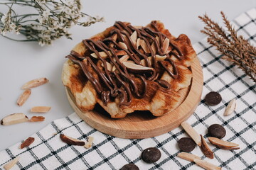 Croissant Waffle Korea pastry. Croffle with almonds and chocolate sauce topping served in wooden plate on square pattern table cloth. Selective focus.