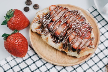 Croissant Waffle or Croffle with chocolate sauce, strawberries and icing topping served in wooden plate on square pattern table cloth. Selective focus.