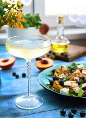 Home brunch atmosphere with a glass of champagne and a fruit salad with grilled peach, feta cheese, blueberry, arugula and olive oil in front of a kitchen window. Blue wooden background