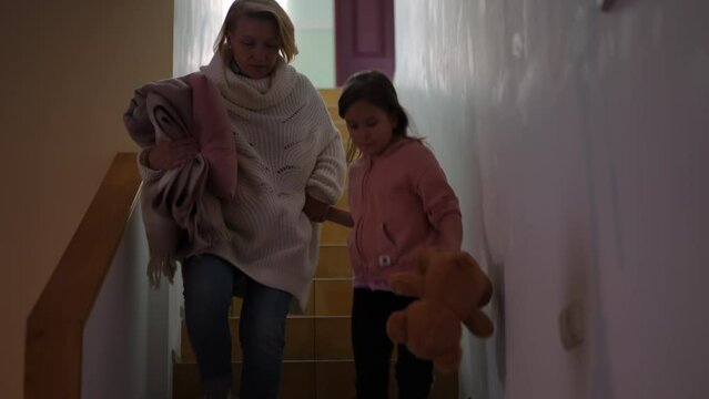 Wide shot anxious woman and girl running downstairs as red air-raid alert warning lamp flashing. Ukrainian worried stressed mother and daughter going to bomb shelter on bombardment
