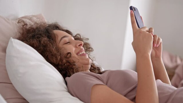 young woman laughing watching funny video lying on bed holding smartphone free morning at home