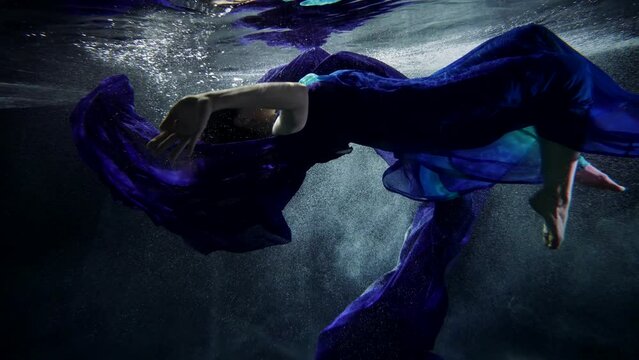 a woman in a blue dress with a long train horizontally hovers face up under the surface of dark water