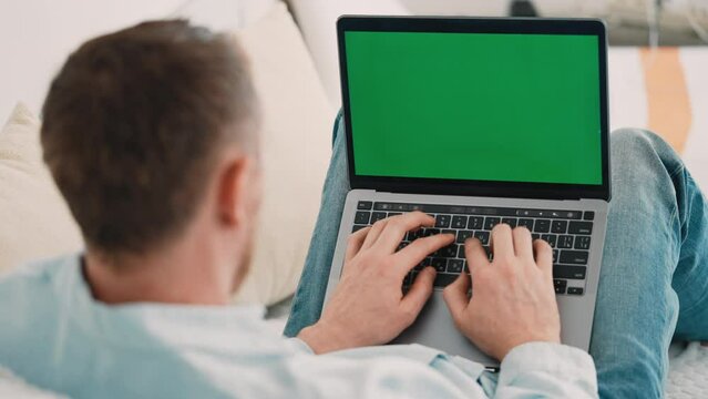 guy typing on laptop green screen displayed sitting on the sofa at home view over the shoulder