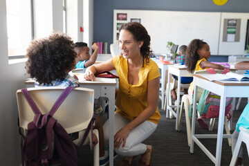 Smiling caucasian young female teacher talking to african american boy sitting at desk in classroom