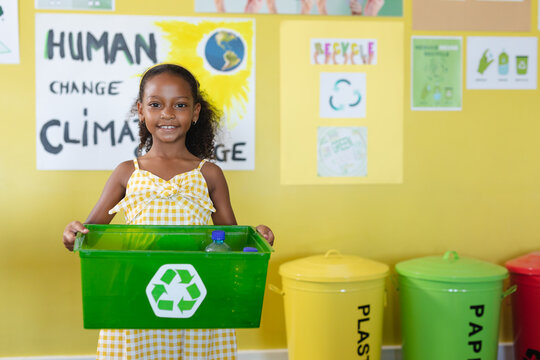 Portrait of smiling african american elementary schoolgirl holding container with recycle symbol