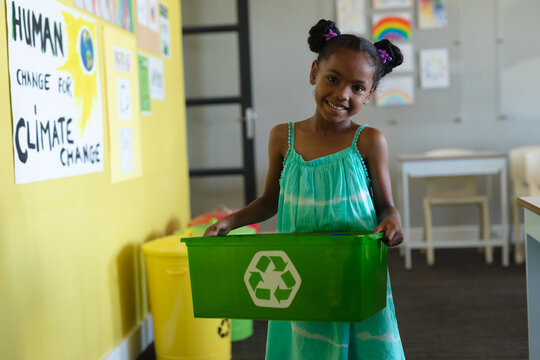 Portrait of smiling african american elementary schoolgirl holding recycle container in school - Powered by Adobe