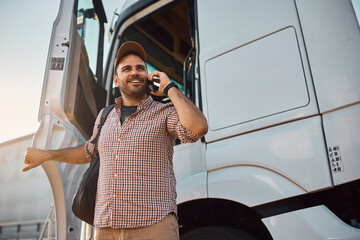 Happy truck driver communicating on mobile phone while getting out of driver's cabin. © Drazen
