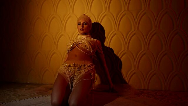 a curvy woman in a turban and an open suit made of shiny beads knelt down and makes a wave against the wall. orange light. the camera is getting closer
