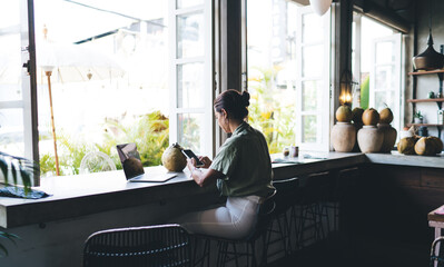 Young millennial woman sitting in cafeteria with smartphone device near laptop computer.Female freelancer using technology and internet for communication. application for social network and banking