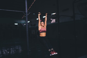Muscular female athlete training body muscles while doing pull ups in gym studio, good looking woman in sportive tracksuit spending morning time for doing workout exercising on horizontal bars