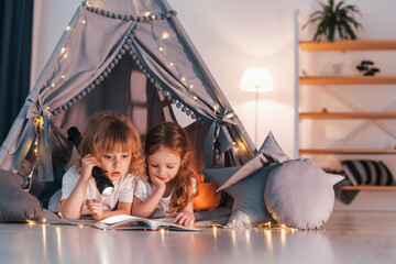 Obraz na płótnie Canvas Reading book and using flashlight. Two little girls is in the tent in domestic room together