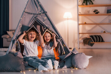 Fototapeta na wymiar Holding books on head. Playful mood. Two little girls is in the tent in domestic room together