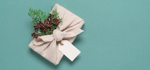 Christmas gift wrapped in fabric with thuja branch, turquoise pastel background. A traditional furoshiki gift. Zero waste concept. Top view, selective focus. Banner