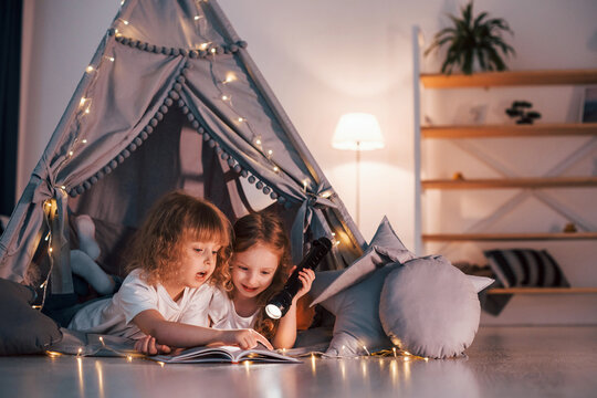 Laying down and using flashlight to read the book. Two little girls is in the tent in domestic room together