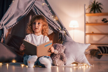 Cute little girl with curly hair is in the tent in domestic room with interesting book