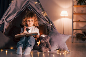 Using flashlight to read the book. Cute little girl is in the tent in domestic room