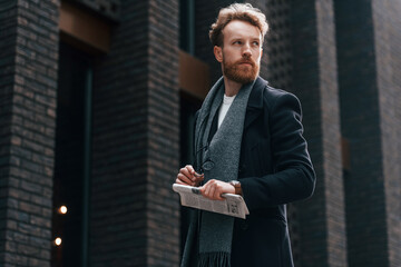 Fototapeta premium Holding newspaper in the hand. Stylish man with beard is outdoors near building