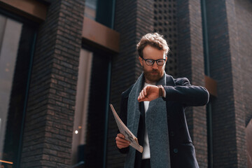 Checking time. Stylish man with beard and in glasses is outdoors near building