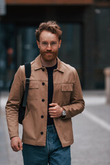 Front view. Stylish man with beard in khaki colored jacket and in jeans is outdoors near building
