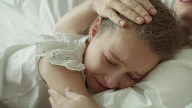 Portrait cute upset child little girl with big tears flowing down his cheeks,child lies at home on bed next to his mother and cries, tears flow with a screaming face.Mom takes pity on her daughter.