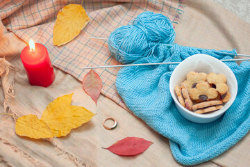 Obraz na płótnie Canvas Tasty cookies in the white plate on the blue yarn with knitting needles, the red candle, the ring and colorful fall leaves. Creativity, leisure, hobby concept background