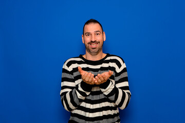 Handsome spanish bearded man in a striped sweater offering something in his hands while looking and smiling at the camera, isolated on blue studio background.