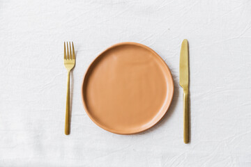 Clay plate of orange color, knife and fork on a white tablecloth of their natural material. Top view