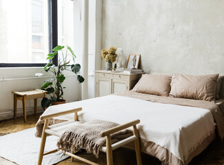 Bohemian styled cozy bedrom with king sized bed and big windows, grey antique furniture by left side of with greenery and and a Greek statue. Bench at the bottom of the bed