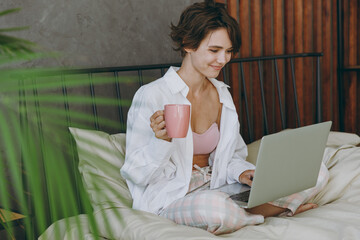 Young student woman wear white shirt pajama she lying in bed use work on laptop pc computer study online hold cup drink coffee rest relax spend time in bedroom lounge home in own room hotel wake up.