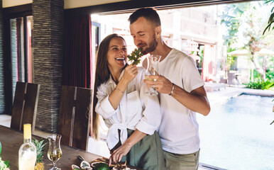 Excited male and female with glass of white wine laughing while cooking brunch meal at house terrace, joyful Caucaisan marriage with food smiling and rejoicing togetherness in homey interior